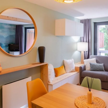 Coin salon avec miroir rond en bois, banquette et coussins moutarde, r&eacute;novation appartement Vars