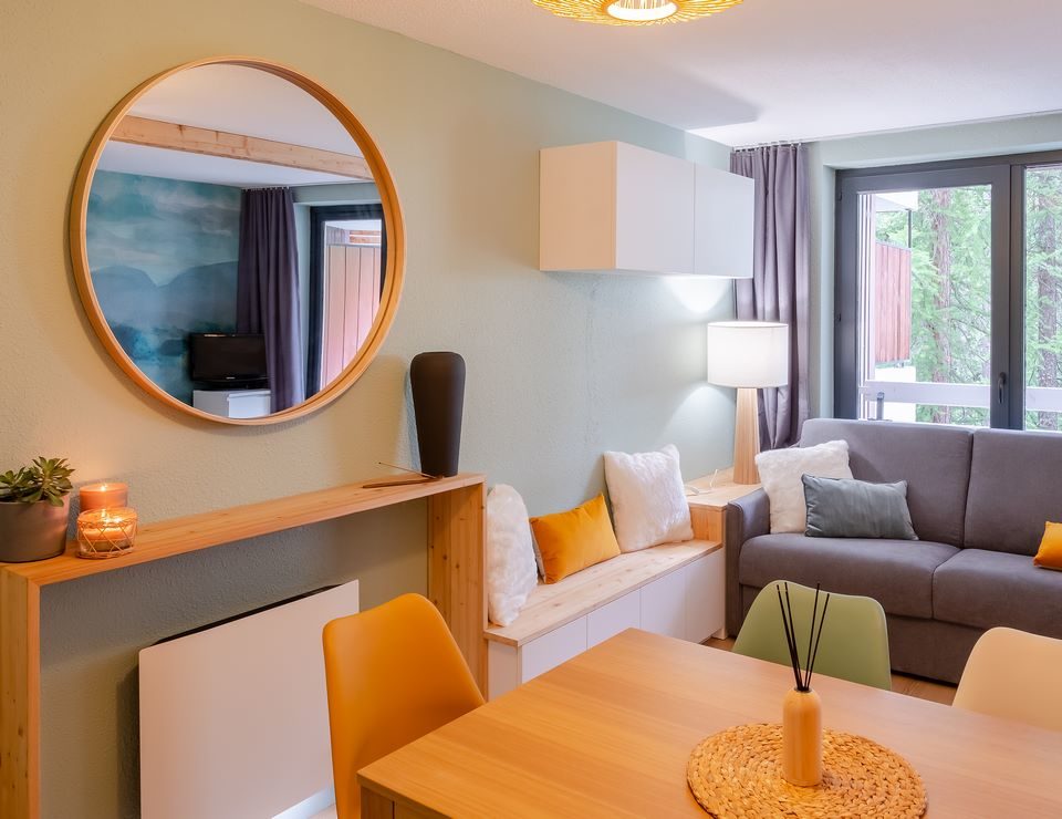 Coin salon avec miroir rond en bois, banquette et coussins moutarde, rénovation appartement Vars